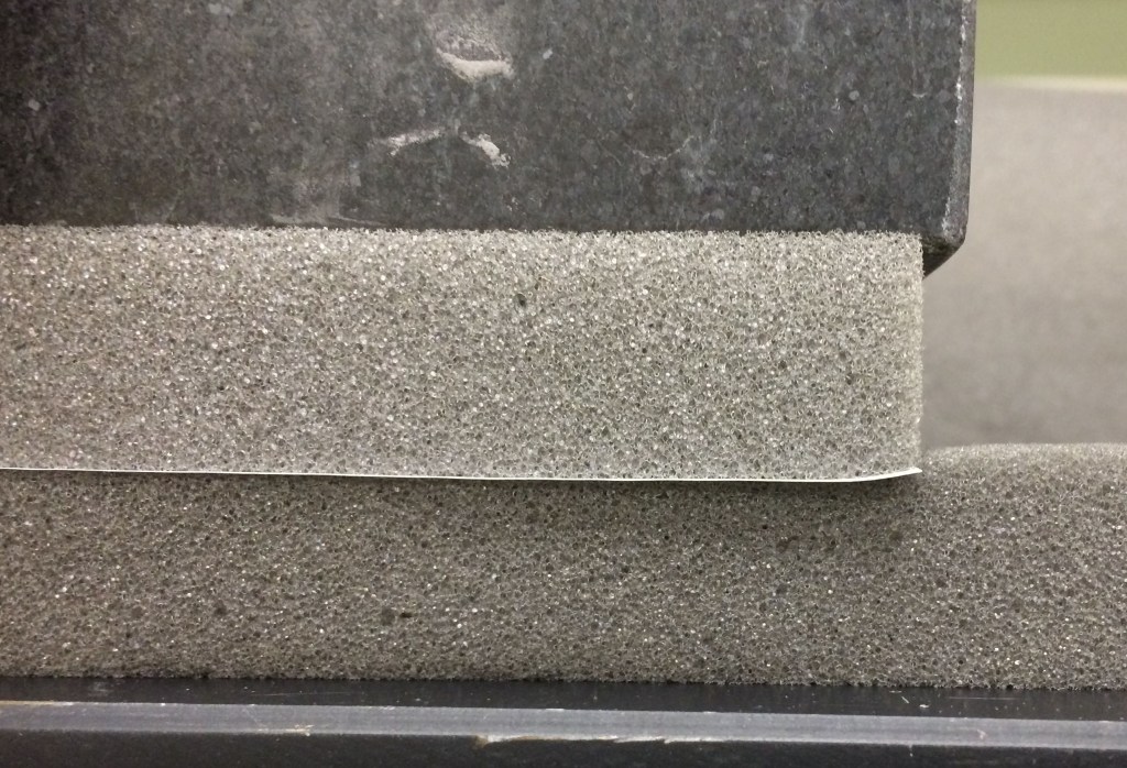 Two pieces of foam compressed by a granite block. The lower piece of foam represents the strop and the upper piece the blade. The interface between these is marked with a sheet of paper.